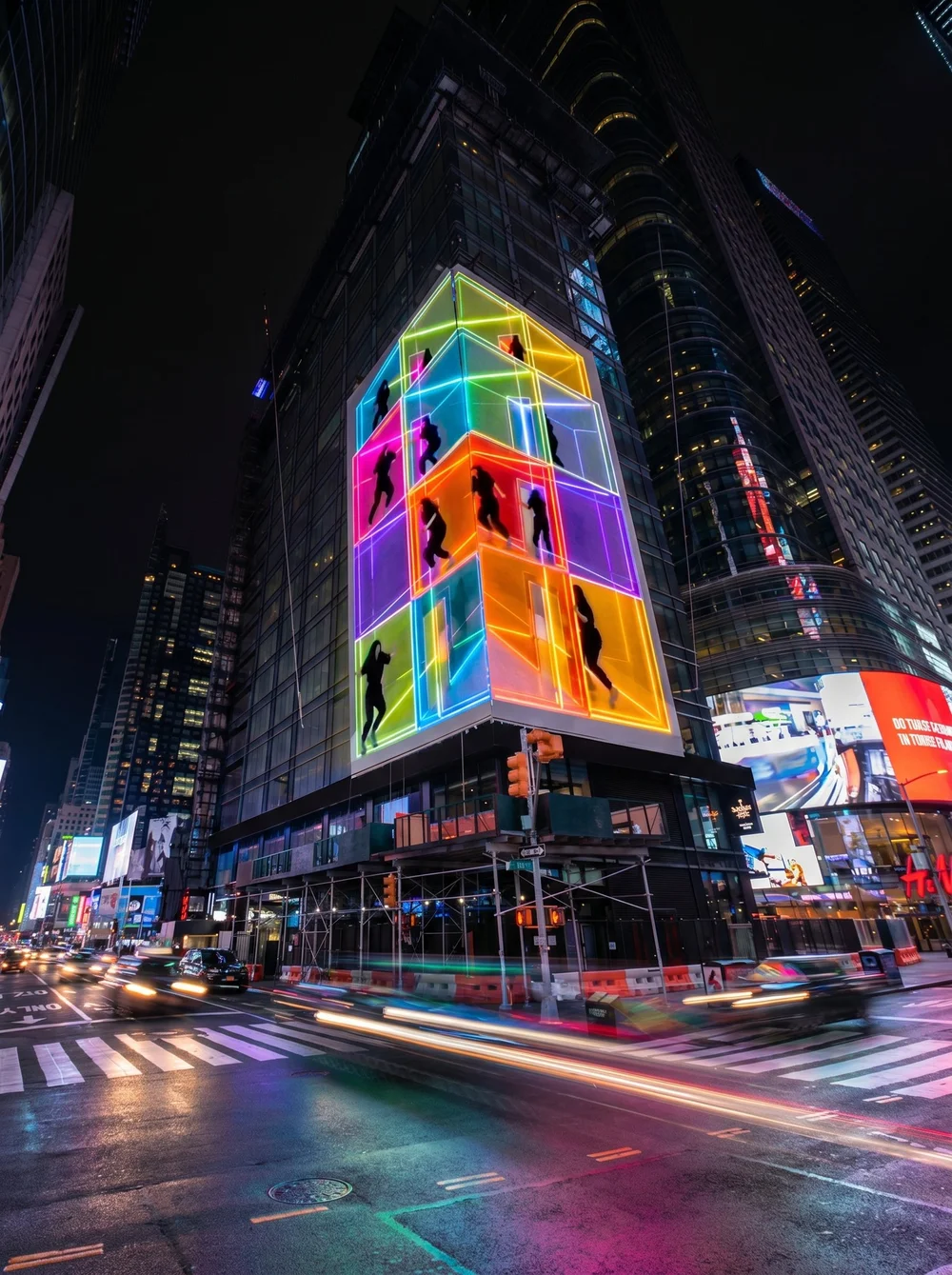 Nighttime city street scene with tall glass building displaying colorful neon light art of silhouettes of people inside defined by bright neon outlines. Traffic moving, creating light streaks on wet road.