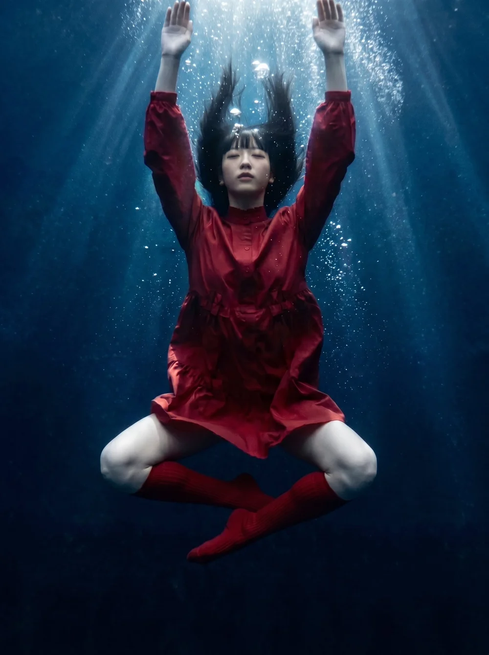 A woman in a red dress, red socks, and red shoes is underwater with her eyes closed, arms raised above her head, and hair floating around her.