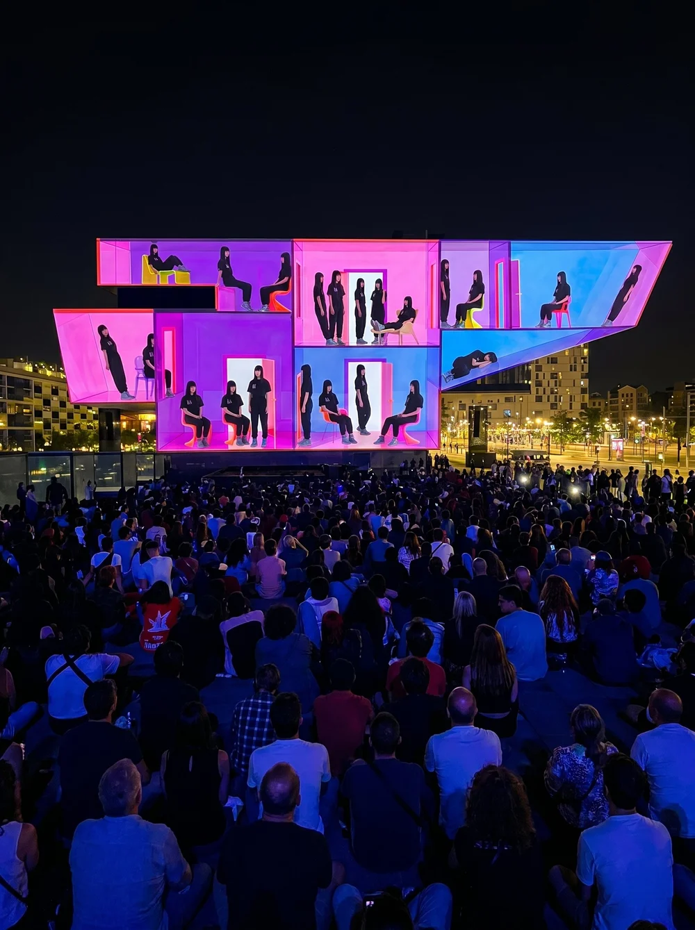 Nighttime outdoor event with a large illuminated modern stage featuring multiple levels and colorful neon lights, displaying numerous people, with audience members seated facing the stage.